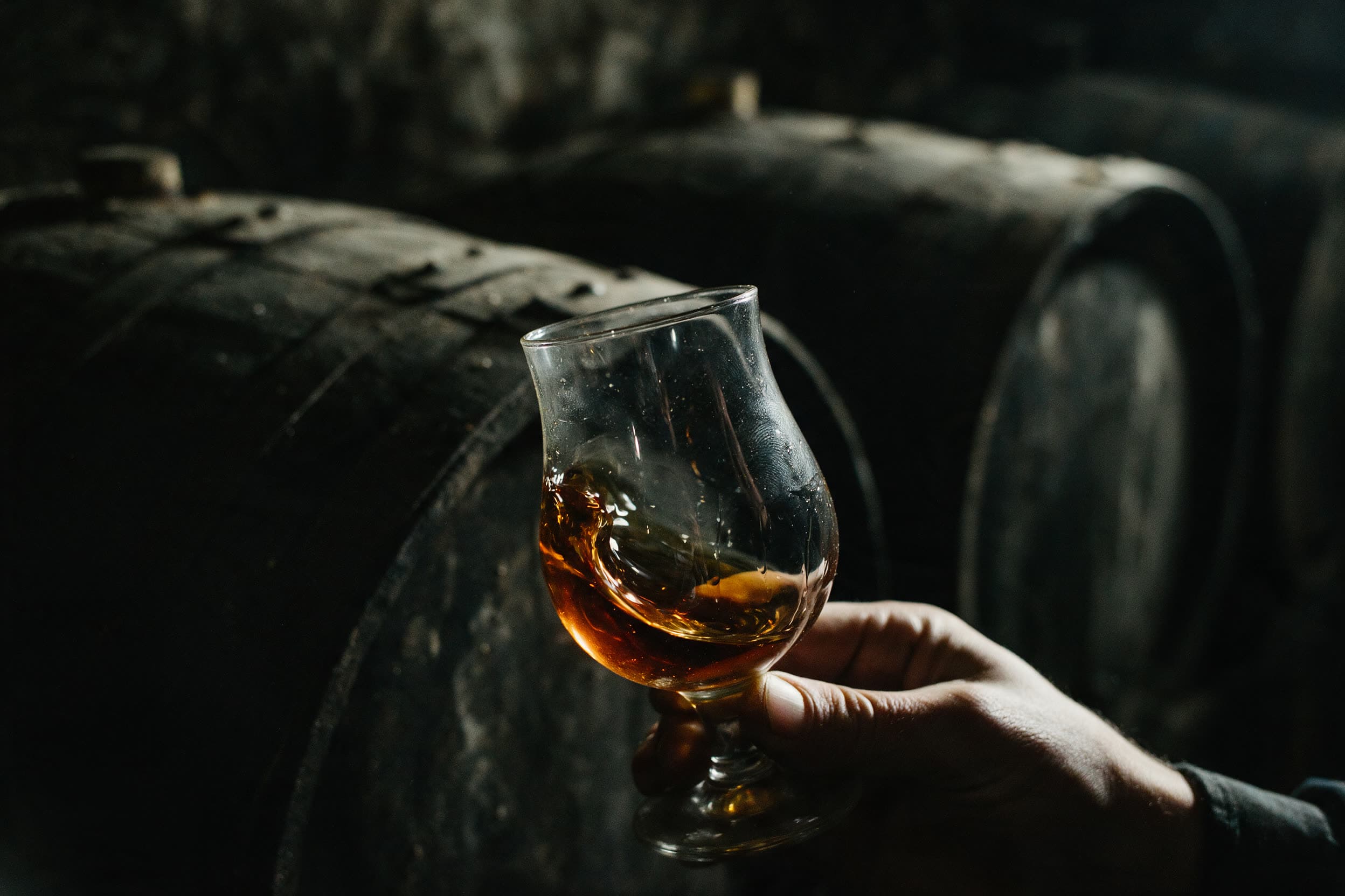 Bernsteinfarbener Brandy wird im Glas geschwenkt, Fässer unscharf dahinter
