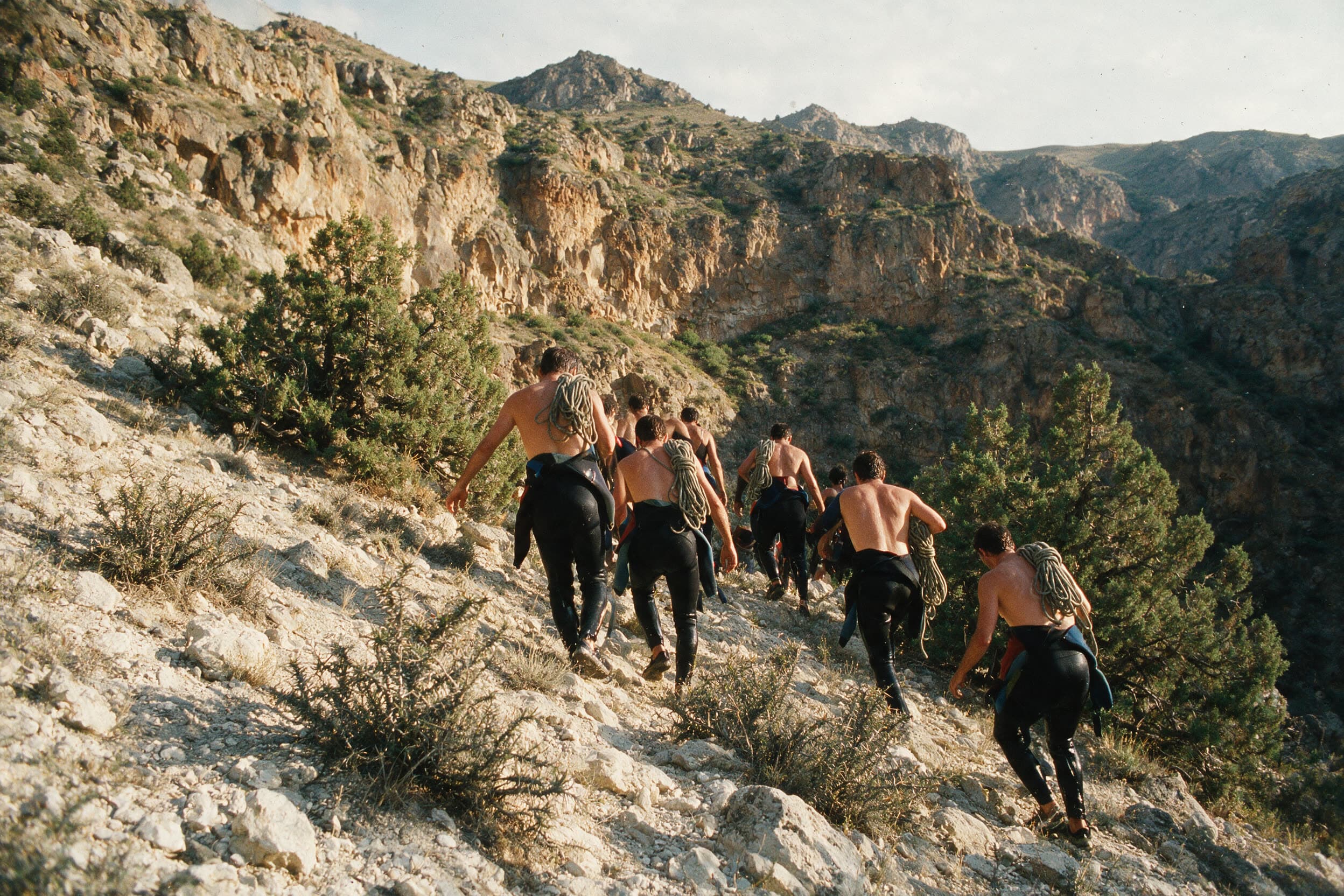 Een groep klimt uit de kloof op een droge zonnige helling, wetsuits half afgestroopt