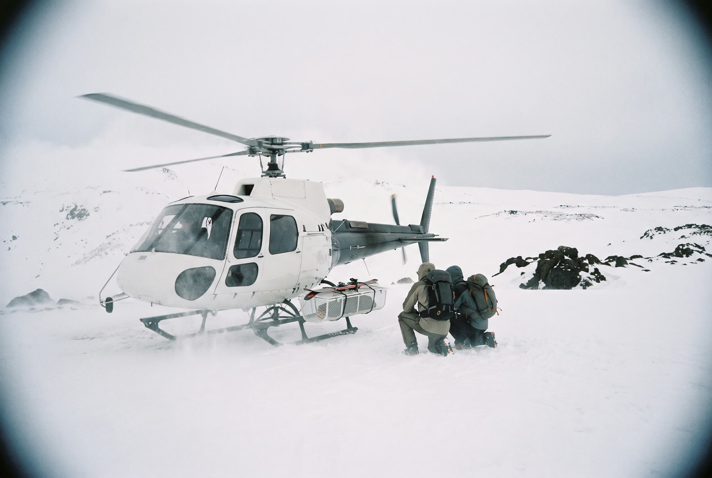 A white helicopter on a snow shoulder of Mount Aragats, two skiers crouching by the skid