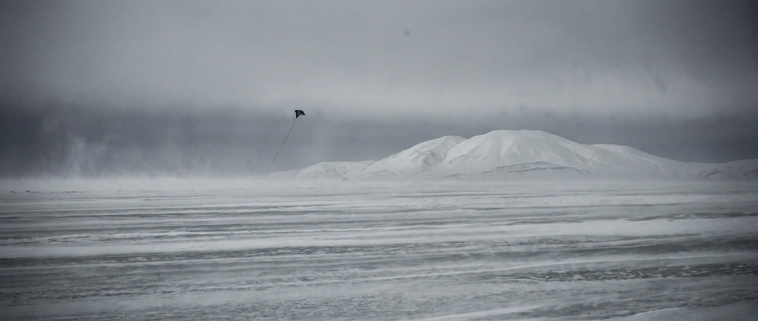 Weite flache Schneehochebene mit Windgangeln, der Kite winzig am grauen Himmel