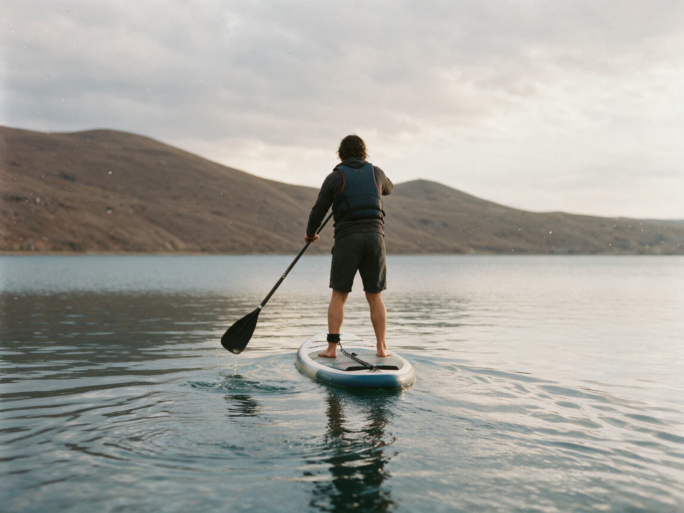 Person auf dem SUP-Board von hinten auf dem stillen Sewansee, kahle Hänge dahinter