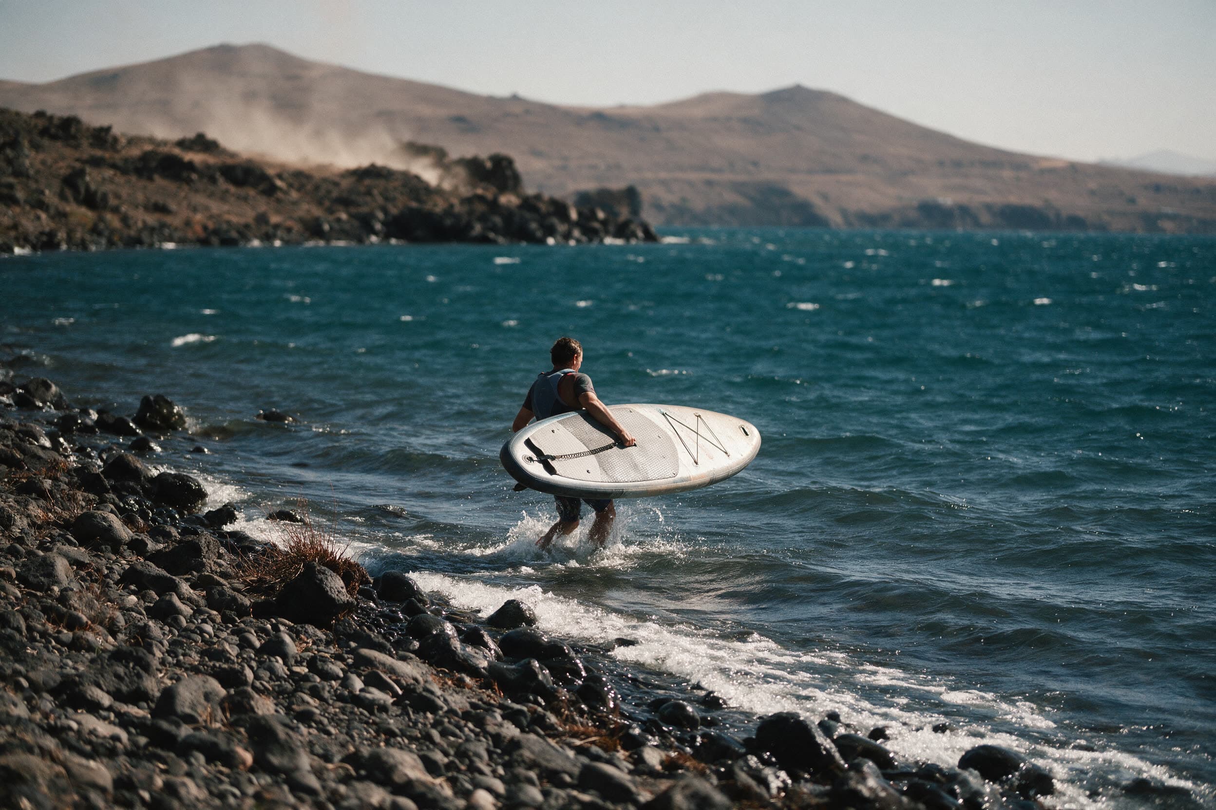 Paddler watet mit dem Board unterm Arm vom felsigen Sewanufer ins Wasser