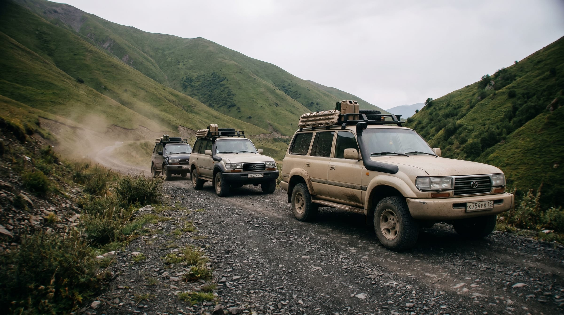 A dusty 4x4 with roof rack and snorkel leads two more along a winding gravel track through green hills.